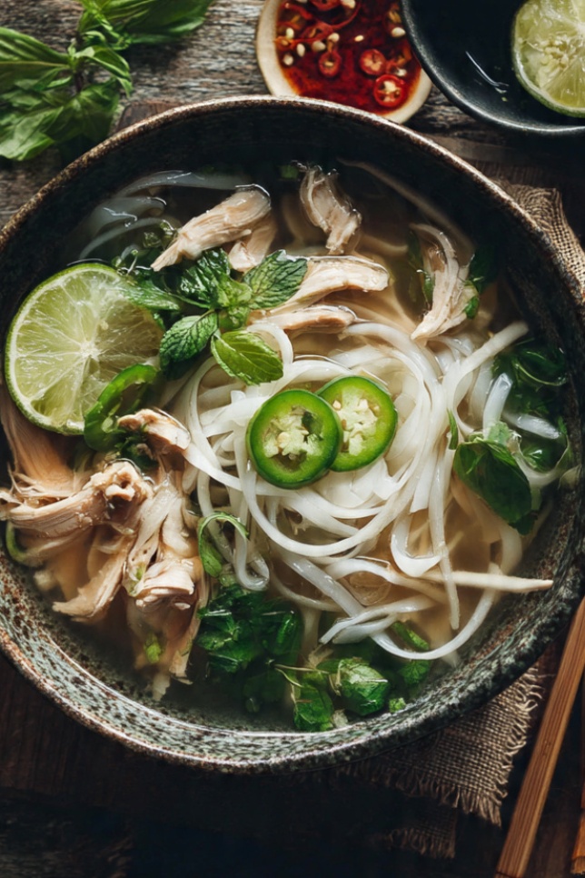 A steaming bowl of Vietnamese pho ga with clear golden broth, tender sliced chicken, silky rice noodles, and fresh Thai basil and cilantro, with lime being squeezed over the top