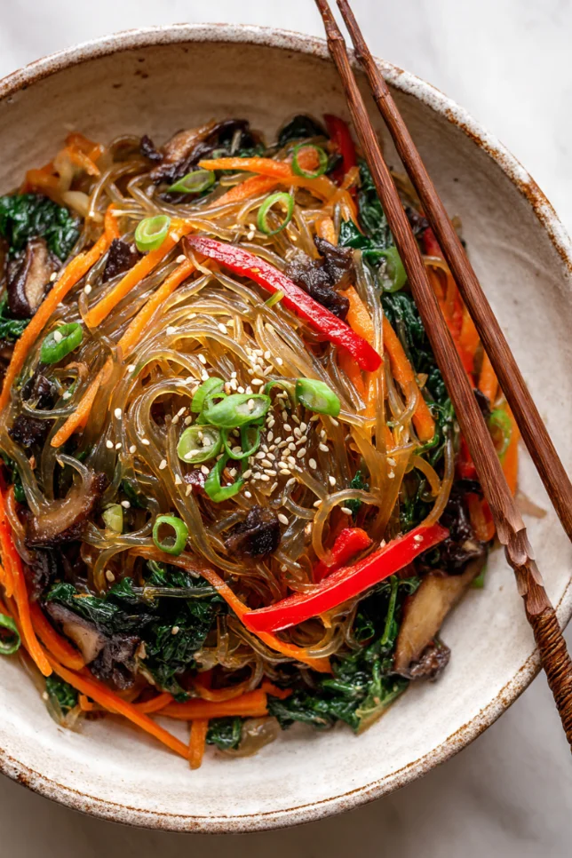 Korean japchae noodles in a white bowl with colorful vegetables, garnished with sesame seeds and green onions