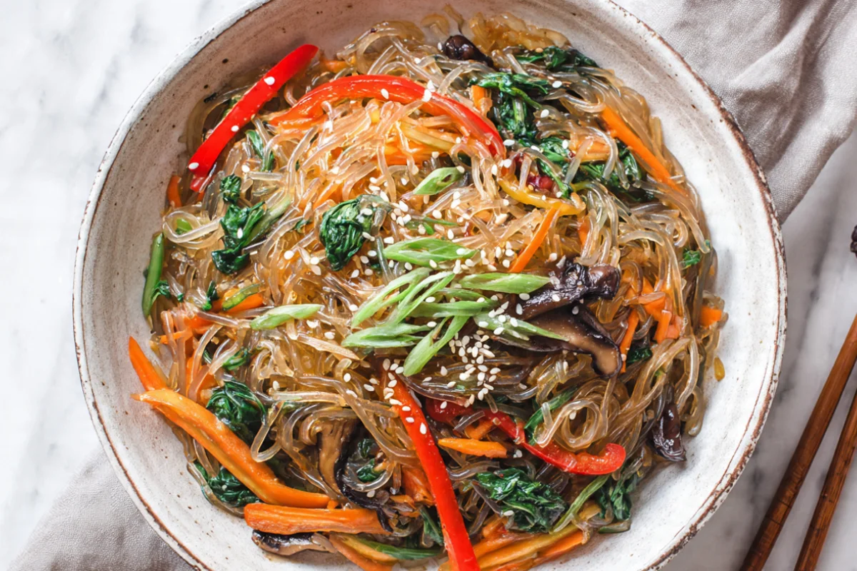 Korean japchae noodles in a white bowl with colorful vegetables, garnished with sesame seeds and green onions